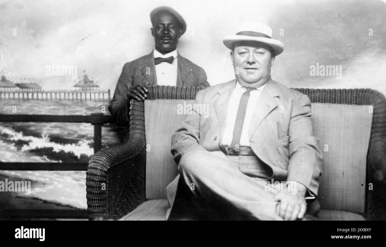 Half length sitting and standing portraits of mature white male and mature African American male, white male wearing light suit with dark tie and hat, African American male wearing dark suit, bow-tie and cap, African American male standing behind chair that the white male is sitting in, standing in front of back drop, neutral expressions, 1920. Stock Photo