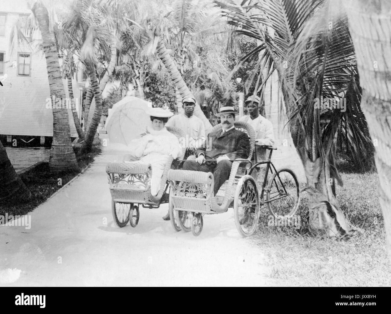 Two African American men carrying white man and woman in single carriages, white woman wearing light dress and light hat with parasol, white male wearing dark suit and hat holding cane, African American men wearing light uniforms and caps, standing and sitting outside in front of palm trees and house, neutral expressions, 1906. Stock Photo