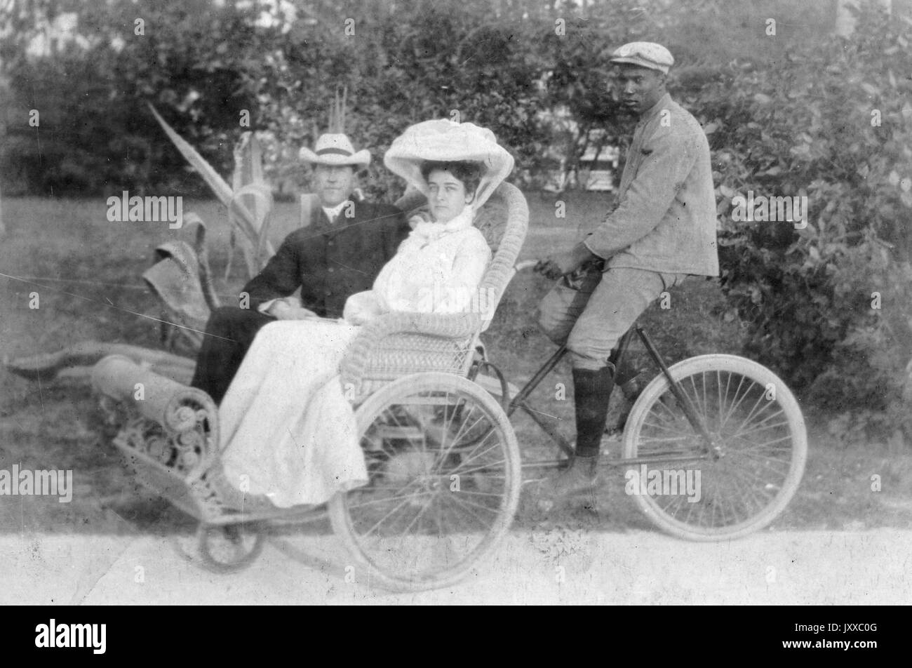African American man bicycling carriage carrying white man and white woman, African American man wearing dark suit with cap, white woman wearing light dress with elaborate hat, white male wearing dark button down shirt, dark pants and hat, standing outdoors in front of vegetation, neutral expressions, 1910. Stock Photo