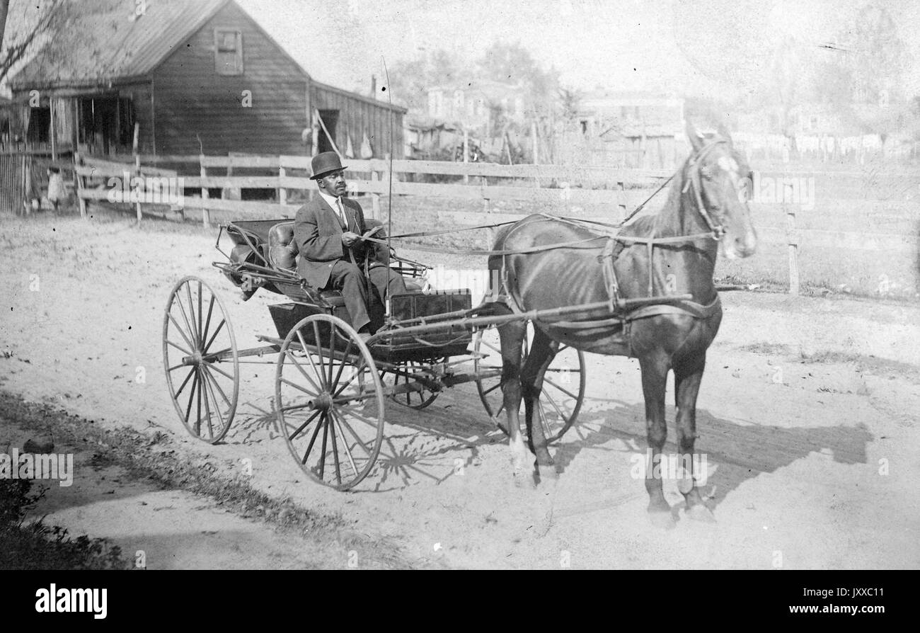 Full length sitting portrait of mature African American male riding horse and buggy, wearing dark suit, light shirt, tie and dark hat, riding along dirt pathway in front of house, neutral expression, 1915. Stock Photo