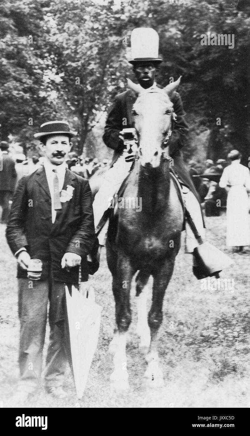 Portrait of African American man riding horse and white man standing, African American man wearing dark jacket, light trousers and hat, white man wearing dark jacket, dark tie, dark trousers and hat, white man carrying umbrella, both outside, neutral expressions, 1920. Stock Photo