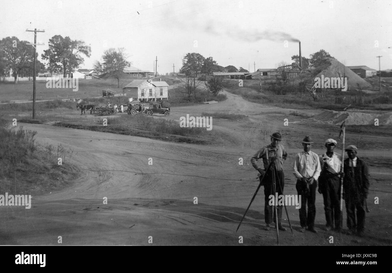 Full length portrait of four men with photography equipment, landscape behind them with a house, horses, mill; surveying team, 1920. Stock Photo