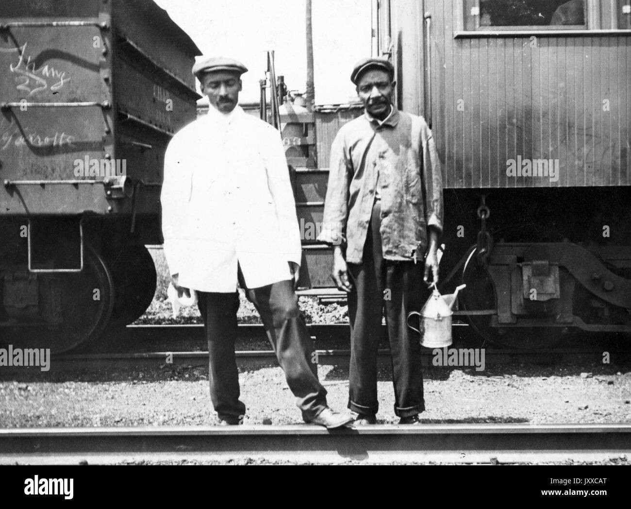 Full length portrait of two African American males dressed casually, one man holding a jug, neutral facial expressions, train behind them; Crew of rear end dynamometer car, 1920. Stock Photo