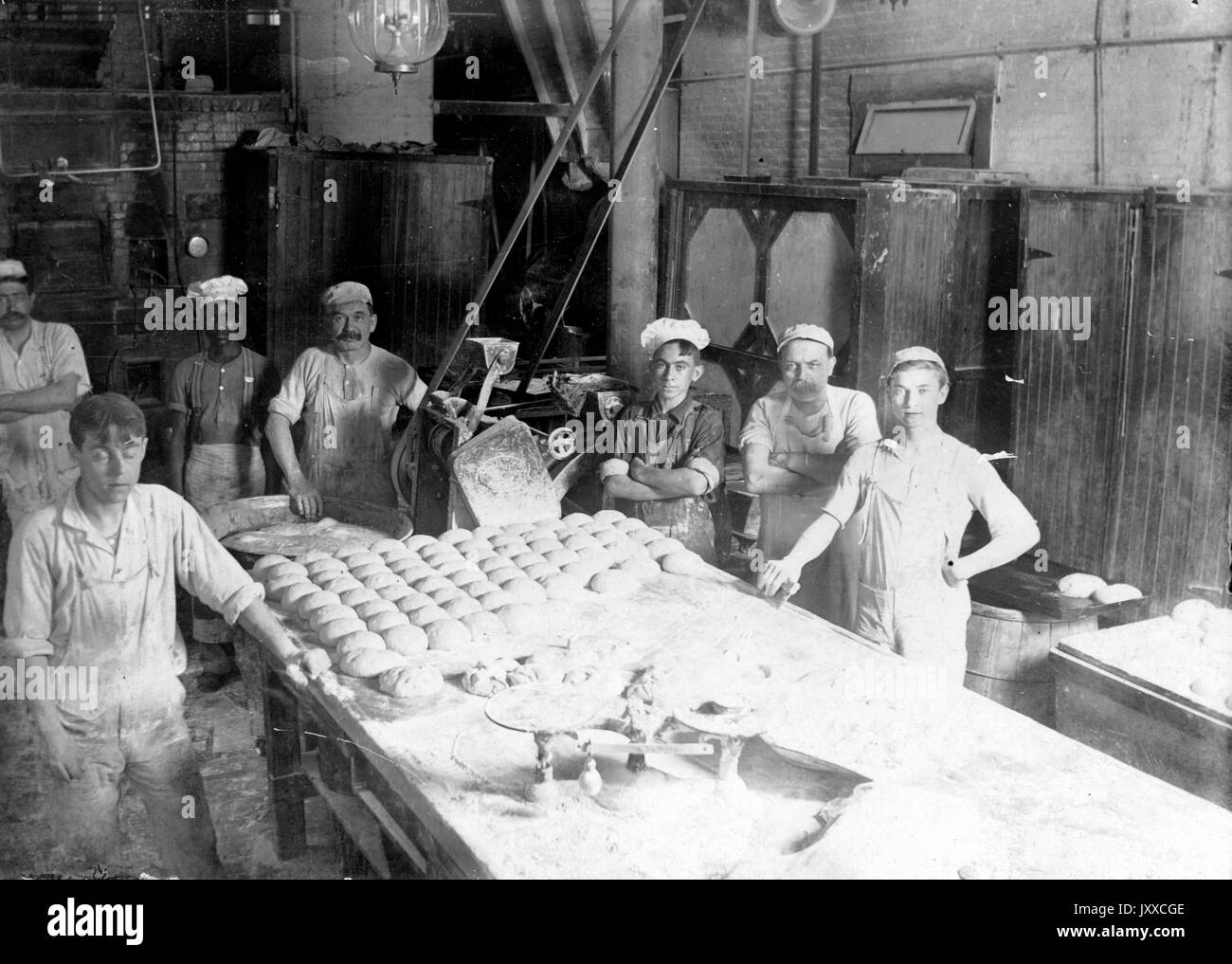 Portrait of seven bakers, standing behind a large table of kneaded dough, working in a brick cellar, wearing white hats, all looking up from their labor, 1920. Stock Photo