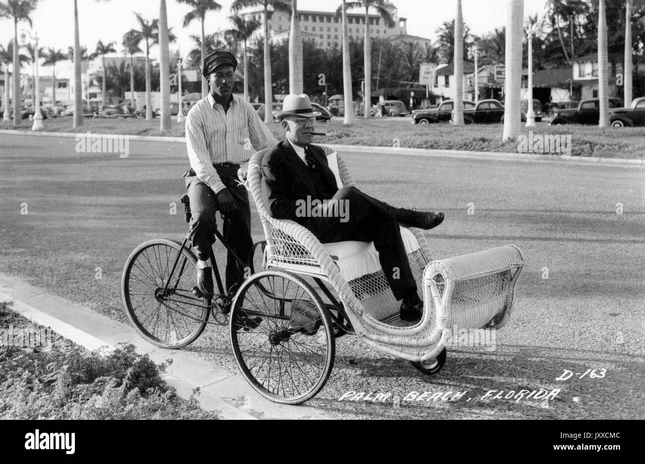 Full Body portrait of an African American man on a rickshaw pushing a mature white man wearing a dark suit white suit and a hat, with a cigar in his mouth, the and African American man is wearing a striped shirt and a dark hat, pushing the man on the grass by palm trees in Palm Beach, Florida, 1929. Stock Photo