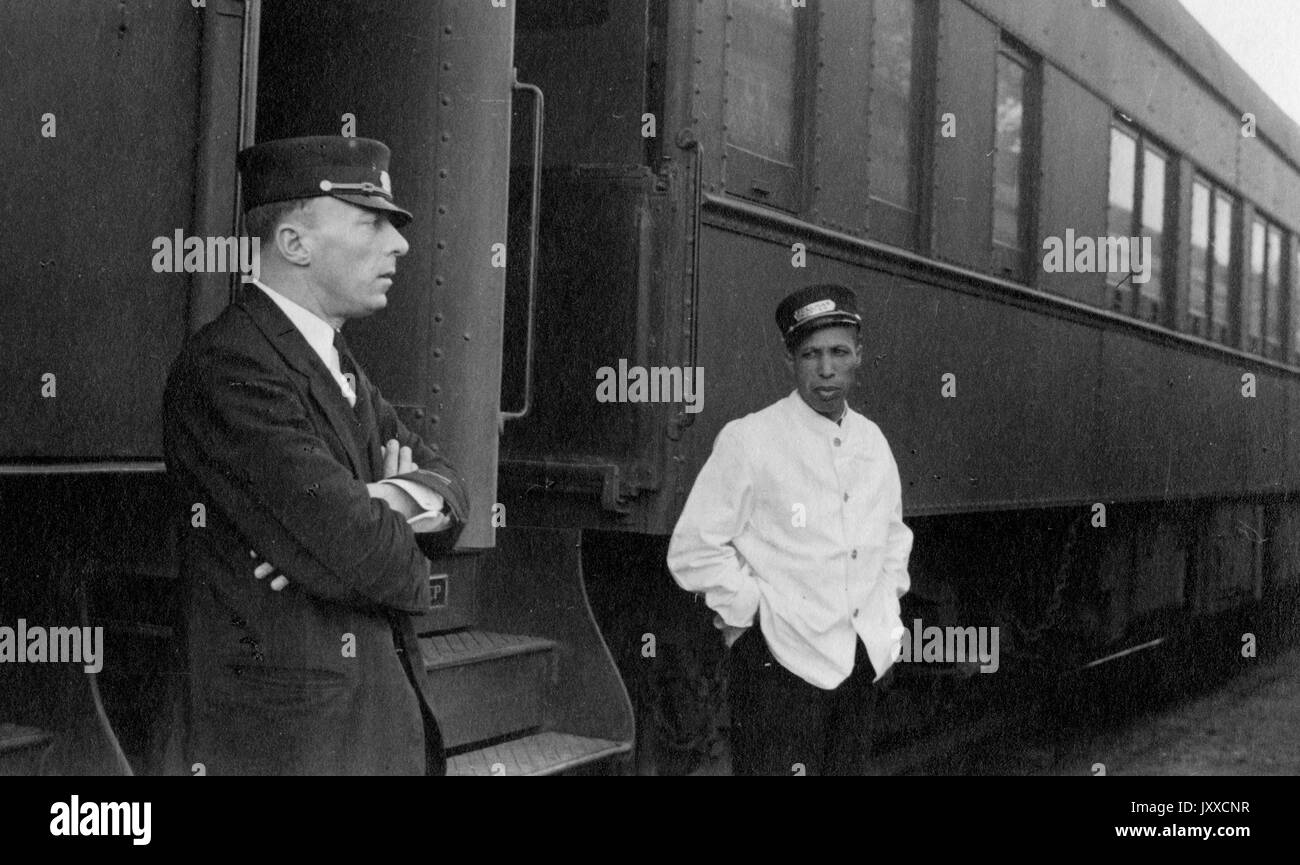 Half-length portrait of two conductors standing outside of a train, one a white man with his arms crossed and wearing a suit, the other an African American man with his hands in his pockets and wearing a white shirt, both donning conductor hats, 1930. Stock Photo