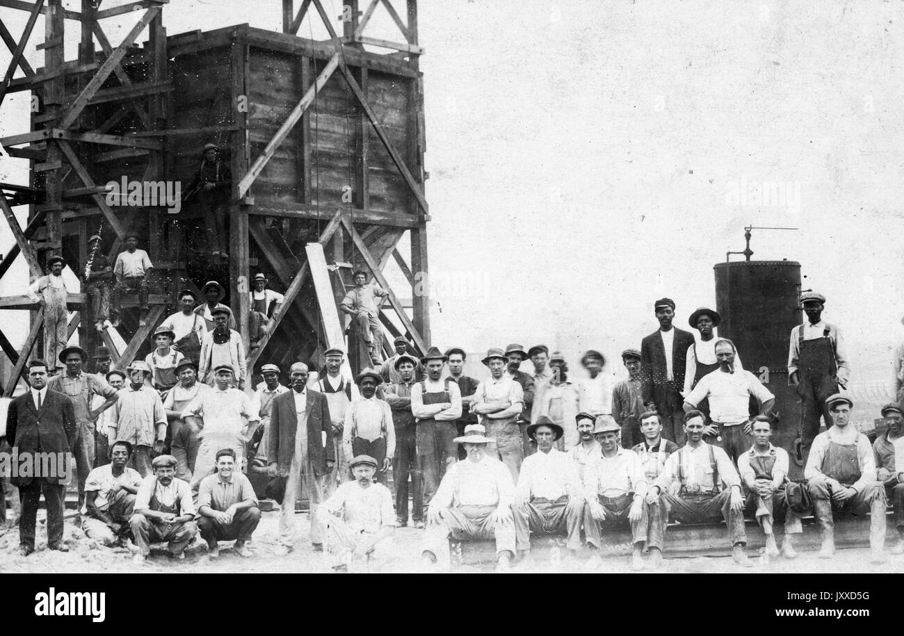 A large group of American workers with neutral expressions is gathered outside in front of, and upon, a large wooden tower structure, 1915. Stock Photo