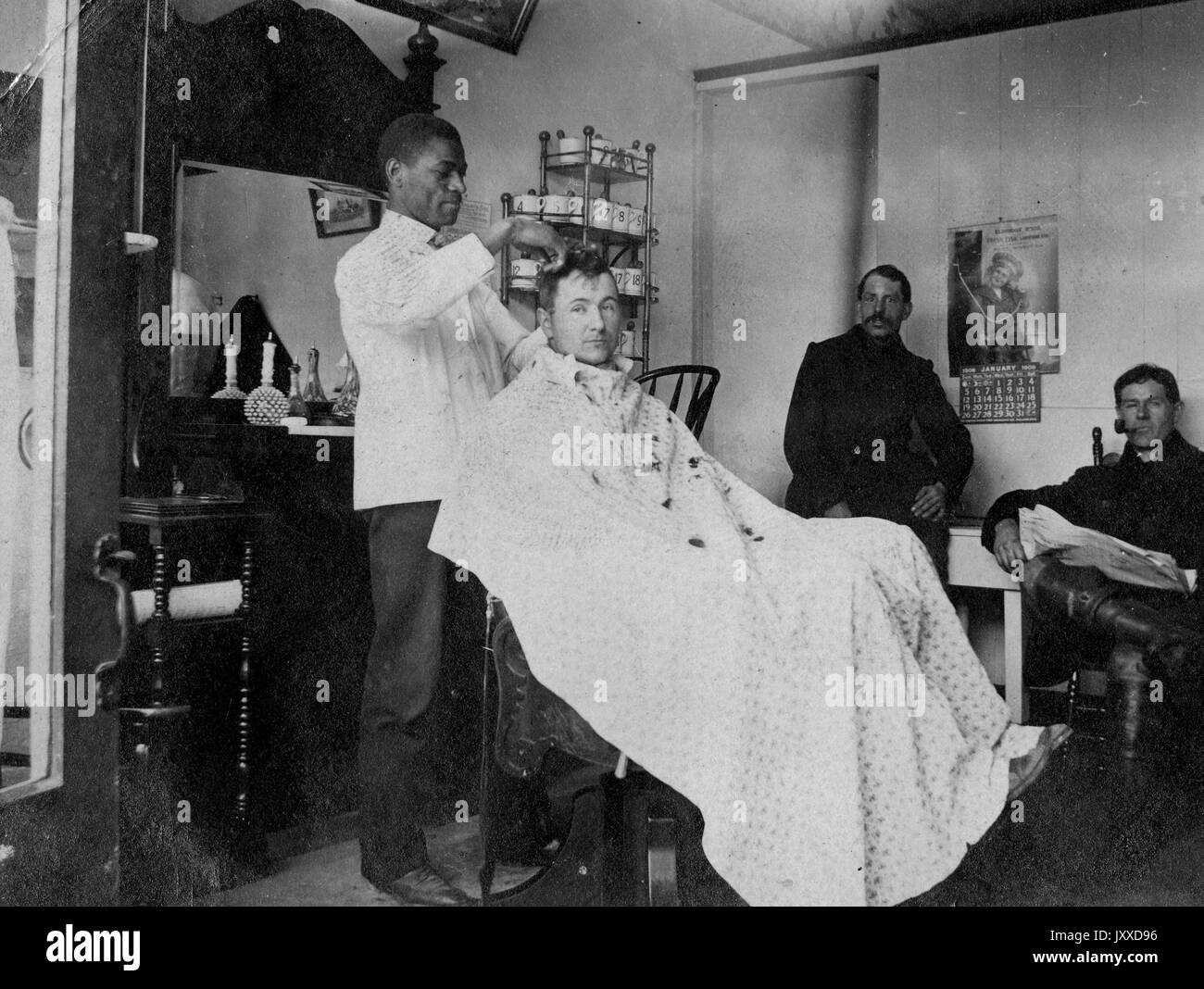A group of mature men with neutral expressions, including one white patron  sitting in a barber chair, one African American barber cutting his hair,  and two white men who stand nearby, are