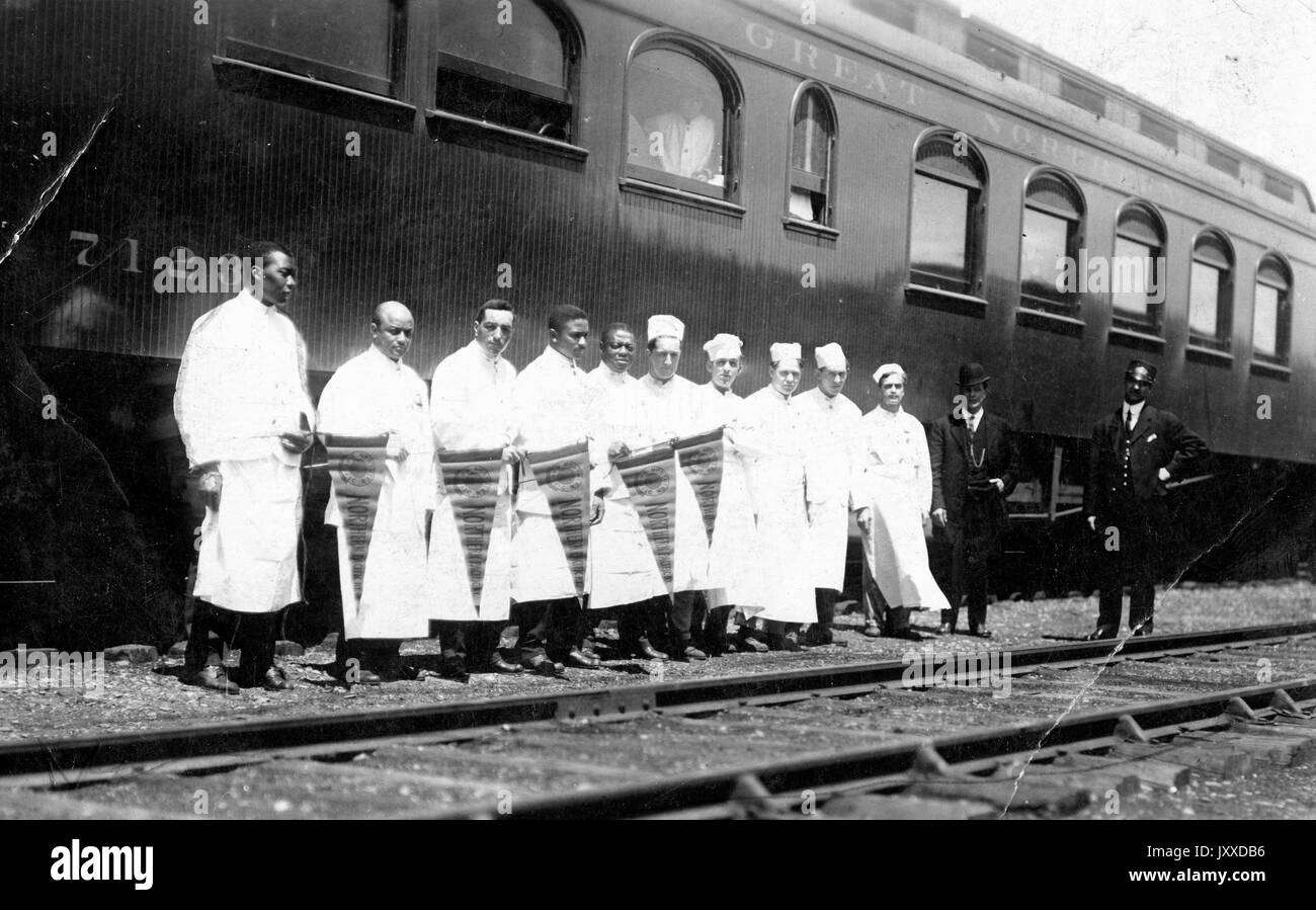 A group of American workers, some dressed in cooks' clothing and carrying pennants, some dressed formally in three piece suits, stand outside by train tracks in front of cart labeled 'Great Northern [Railroad]', 1915. Stock Photo