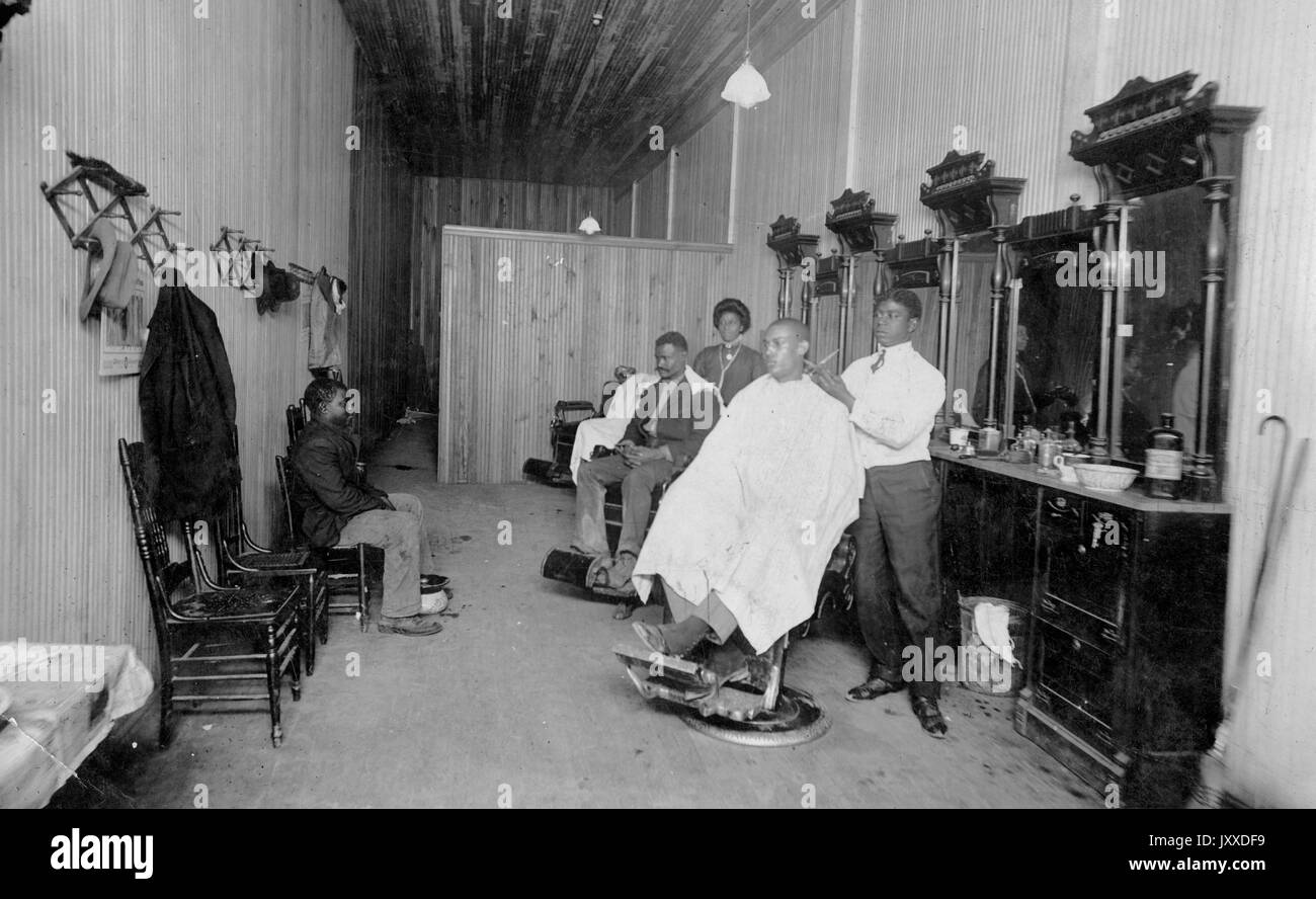 A group of African Americans, including two male patrons, one young man waiting in a chair, one young male barber, and one woman, are gathered in a barbershop, containing chairs, hat and coat racks, and three stations with barbers' chairs, mirrors, and equipment, 1912. Stock Photo