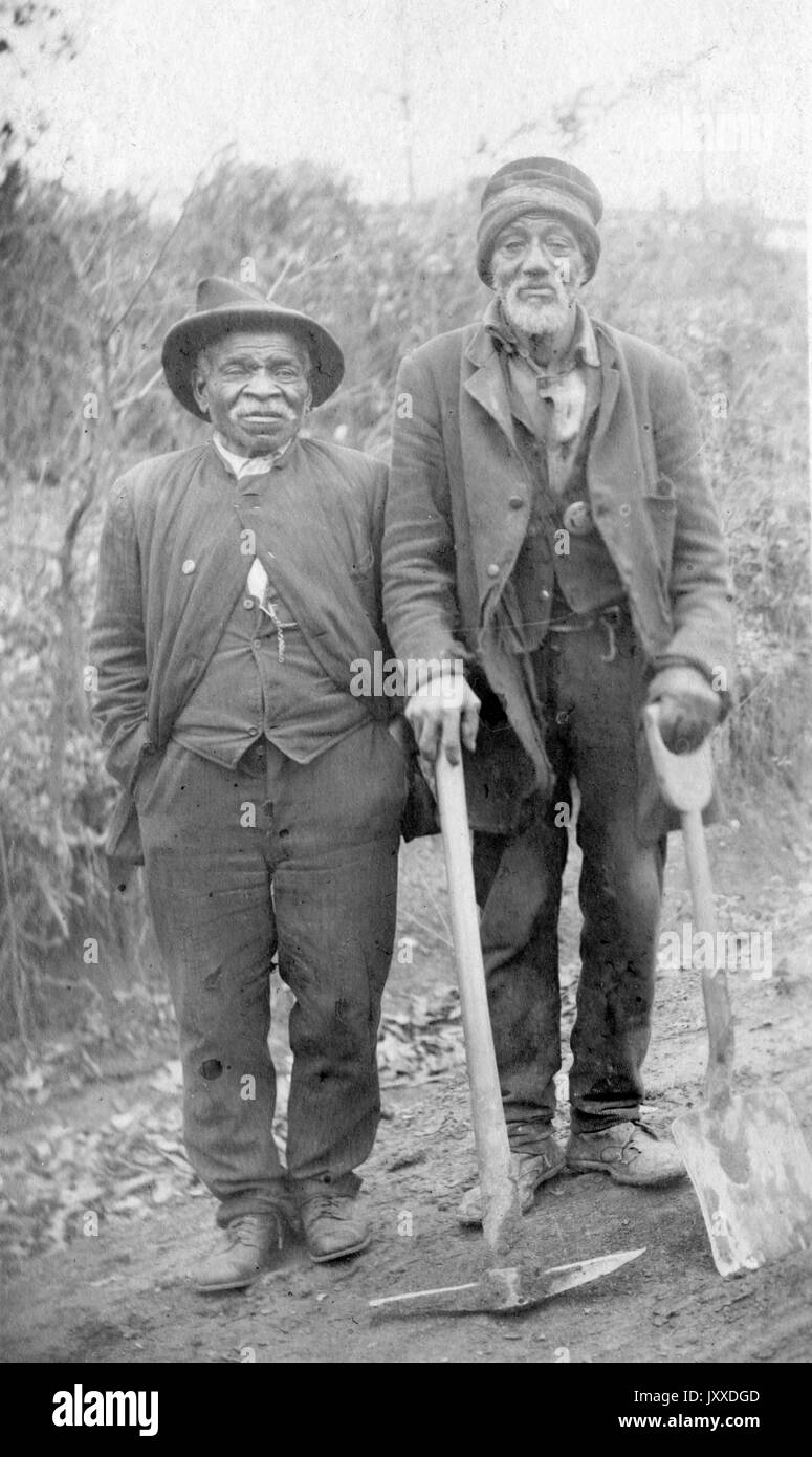 Full length standing portrait of two mature African American workers with neutral expressions standing in a cleared wooded area with a pickax and a shovel, 1920. Stock Photo