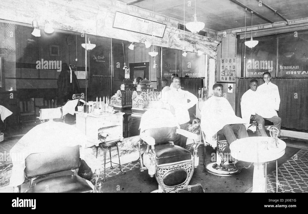 Two African American men sit in barber chairs, each with an African American barbers standing by, in a barbershop with a patterned floor, many lights, and marble accents, 1920. Stock Photo