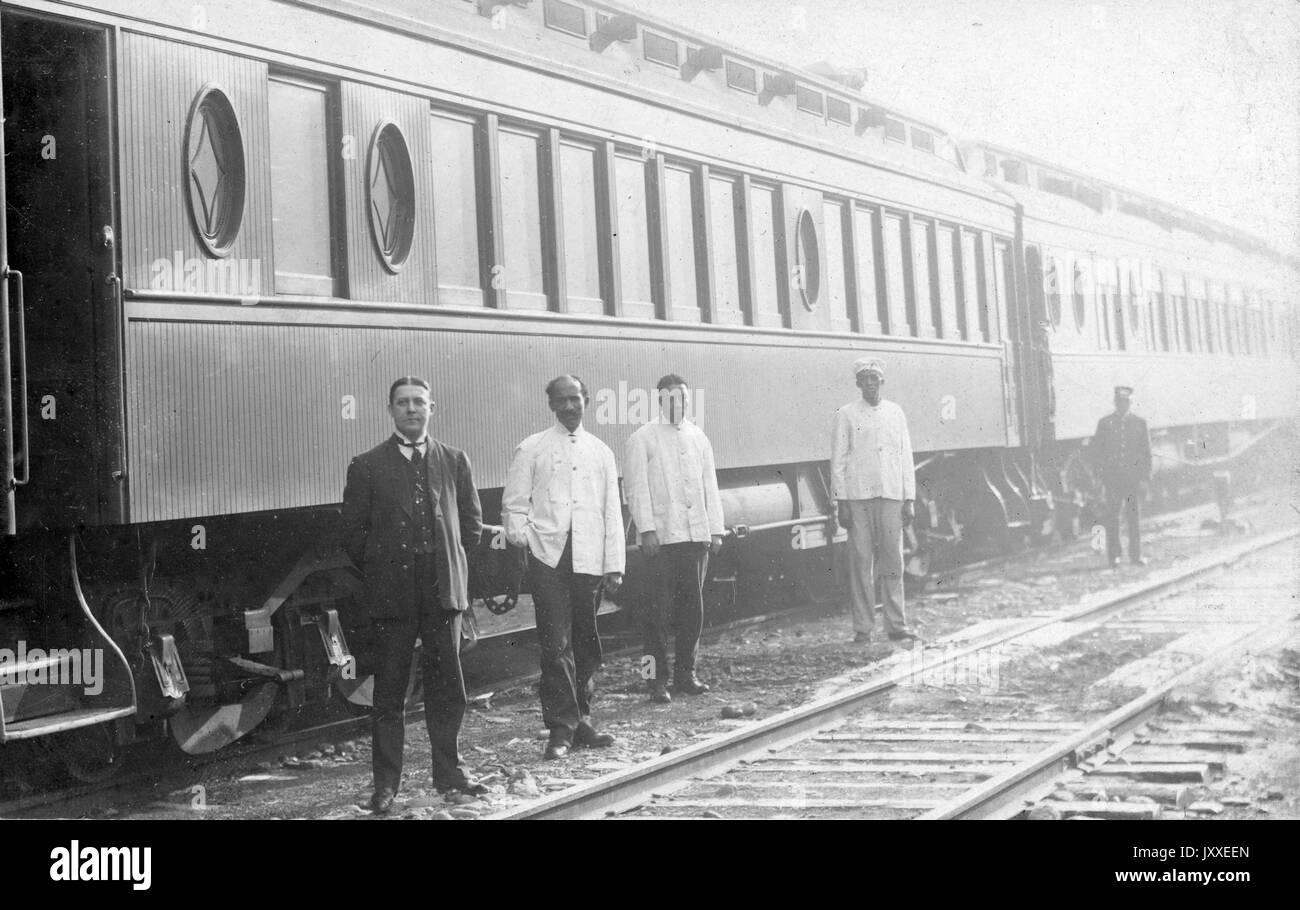 African American train employees are standing next to a white train employee in front of a train, the African American men are wearing light colored shirts and dark pants and the white employee is wearing a full dark colored uniform, 1920. Stock Photo