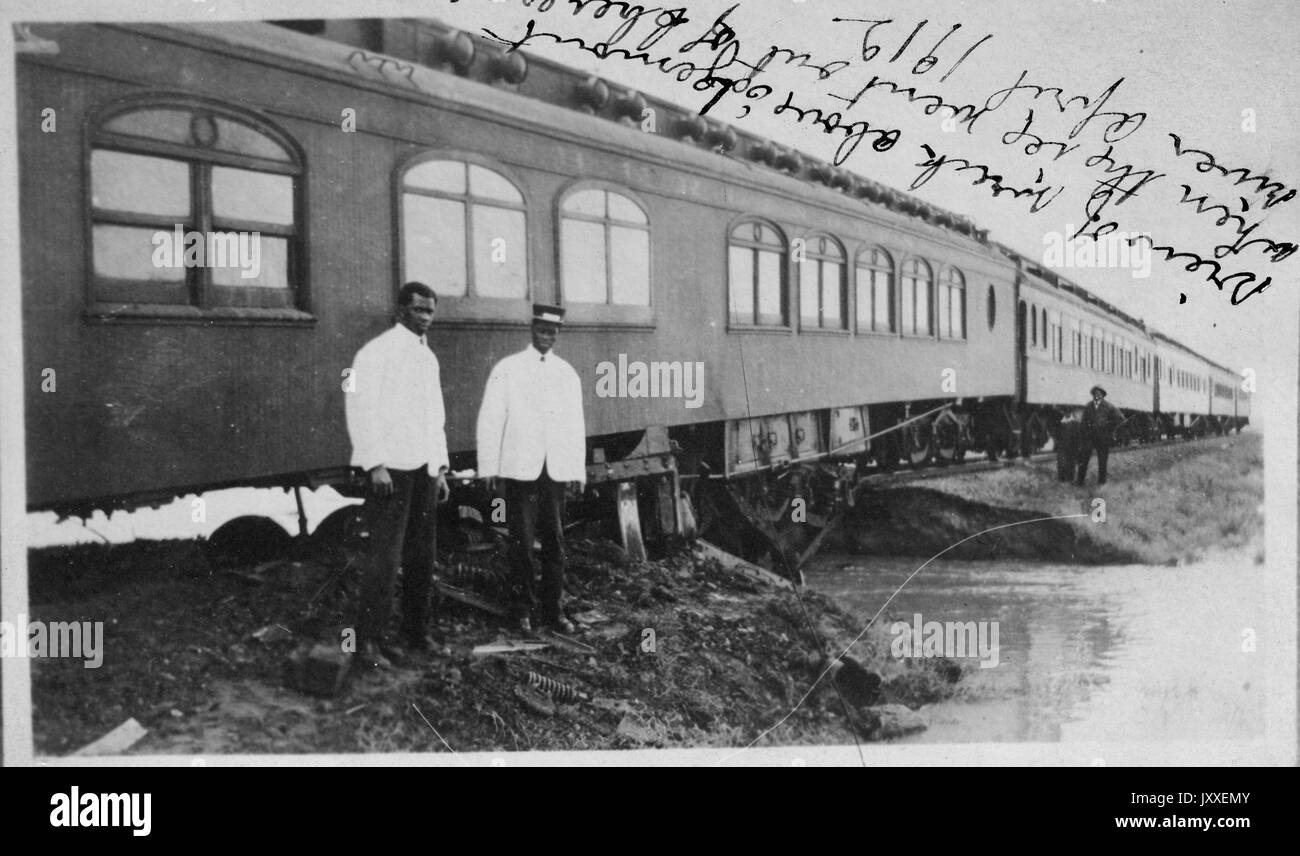 Two African American train employees standing in front of a train, the train is crossing over a small bridge with water underneath, a railroad worker is on the other side of the patch of water, the two employees are wearing light colored shirts and dark colored pants, 1912. Stock Photo