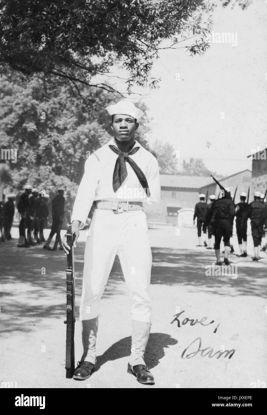 Portrait of an African American US Navy Sailor standing in front of two groups of sailors practicing in dark colored outfits, he is wearing a light colored Sailor uniform, one hand is behind his back and the other is holding up a firearm by his side, 1920. Stock Photo