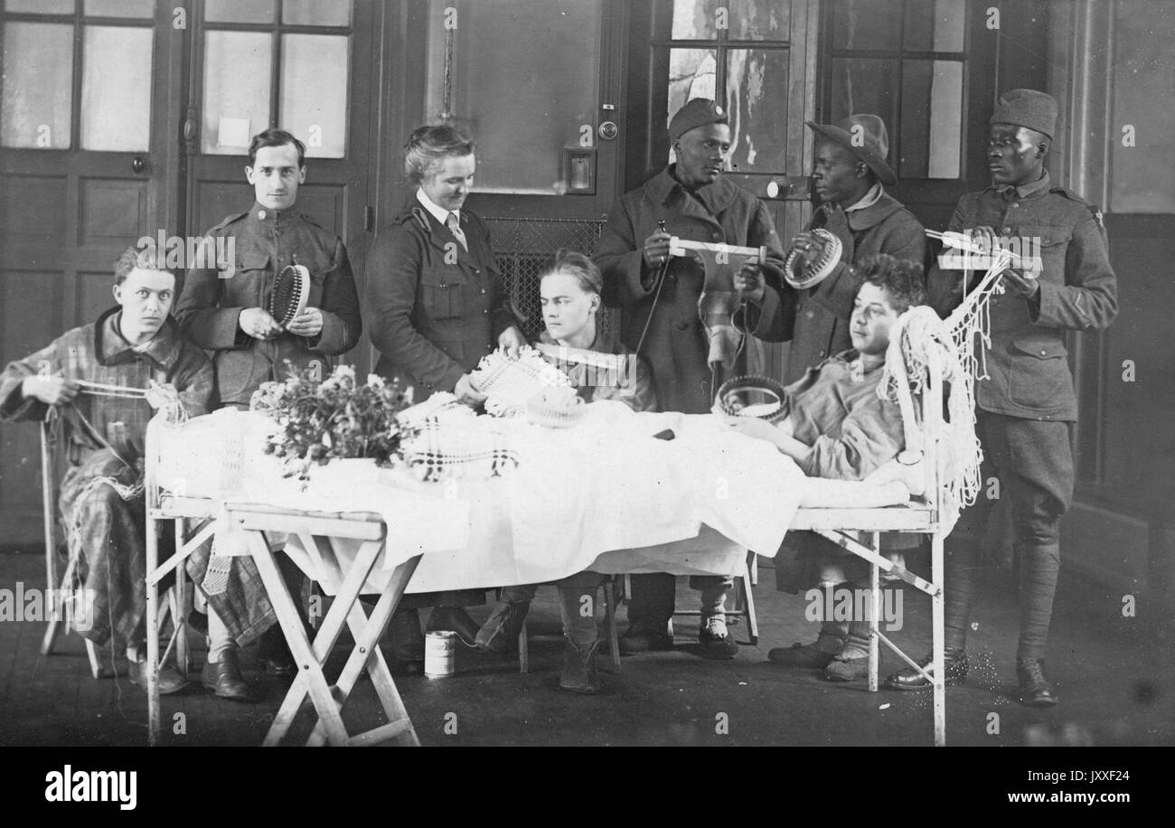 One white young man is perched up while his body lays on a long bed, there are other young white men sitting and standing near him holding knitting supplies, three African American men are standing behind the young man and are holding knitting supplies, a woman is instructing one of the young white men on what to do, 1920. Stock Photo