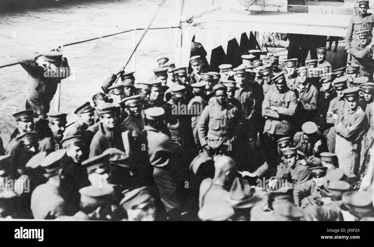 African American and white United States soldiers are aboard a boat on the sea, they are all in uniform and standing around and listening to a solider who is talking, 1920. Stock Photo