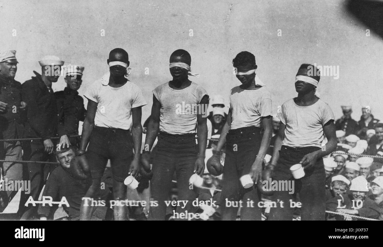 Four African American boys are standing in a boxing ring with blindfolds on, they have a boxing glove on one hand and a mug in the other hand, hey are wearing light colored t-shirts and dark pans, behind them are white naval soldiers dressed in uniform and looking on, 1920. Stock Photo