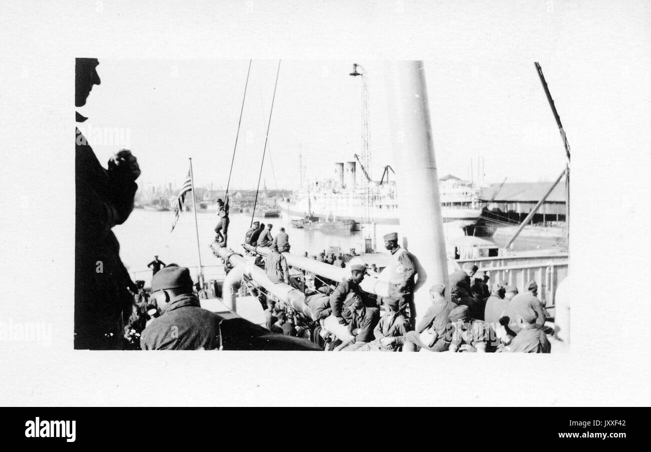 African American and white naval soldiers are siting on a large ship docked at a port, in the background is another ship and more of the port, 1920. Stock Photo