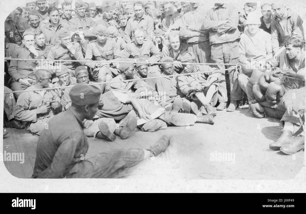 A large group of African American and white soldiers in uniform sitting and standing around an African American soldier sitting on the ground, the soldier is sitting with his legs stretched out and crossed at the knees, there is a rope separating the solder from the group of onlookers, there is a pile of boxing gloves towards the right near some white soldiers, 1920. Stock Photo