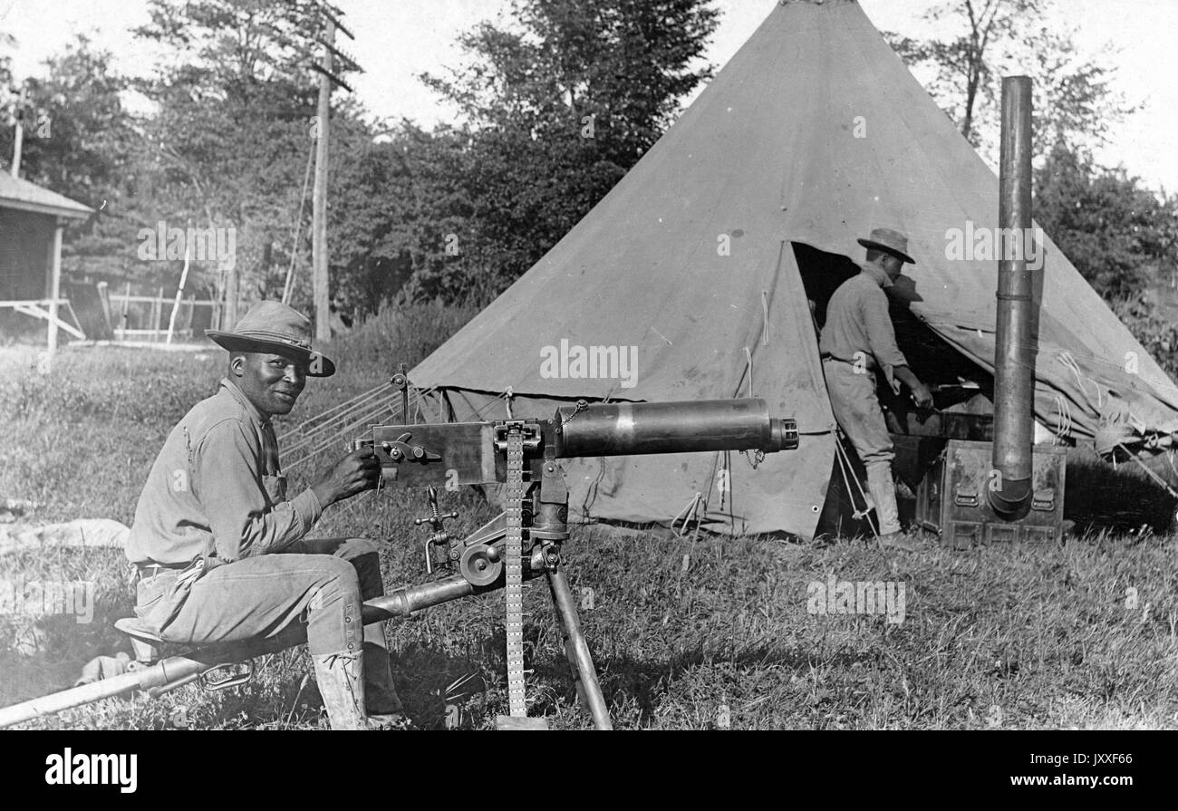 Two African American soldiers are by their tent, one is sitting with a piece of military equipment and the other is half inside of the tent standing, there are trees in the background, 1920. Stock Photo