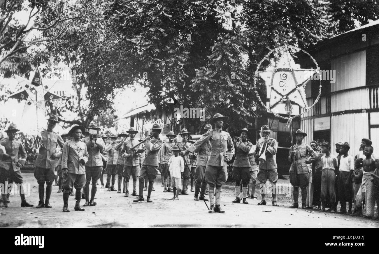 African American soldiers along with some non-army members are marching in a parade, most of the army men are playing instruments, there are two larger star signs, one star has the number 19 on it, there are African American boys looking on, 1920. Stock Photo