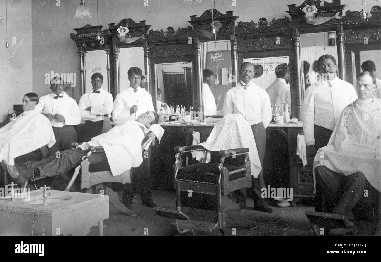 Landscape shot of barbershop, four African American barbers, three customers, wardrobes with mirrors behind the barbers, neutral facial expressions, 1920. Stock Photo
