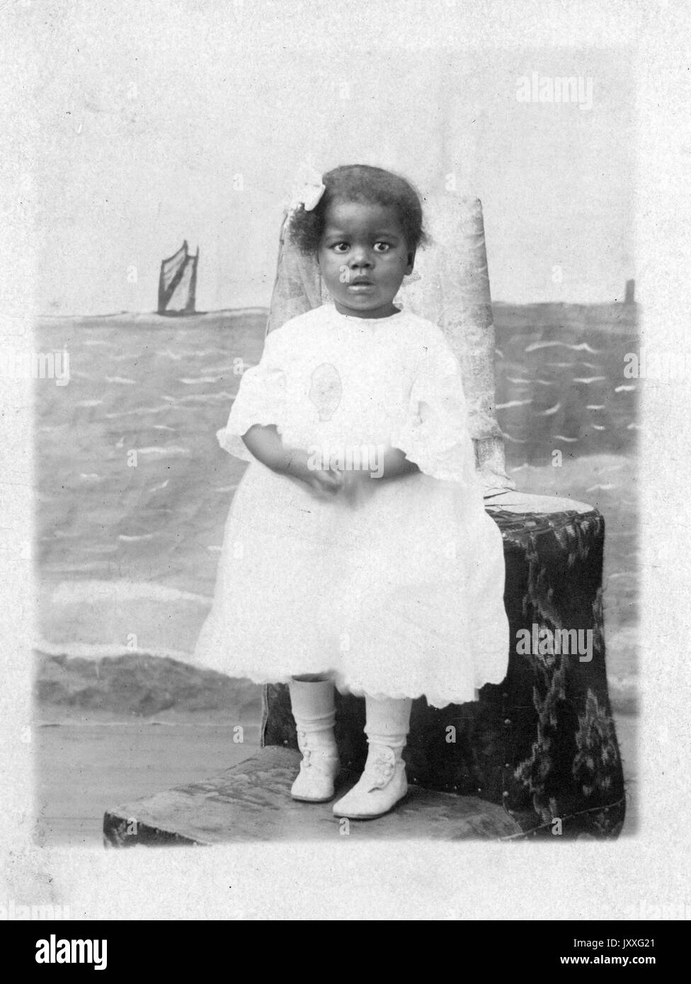 Full length portrait of an African American female child, wearing a white dress, with white shoes, in a photography studio, neutral facial expression, 1920. Stock Photo