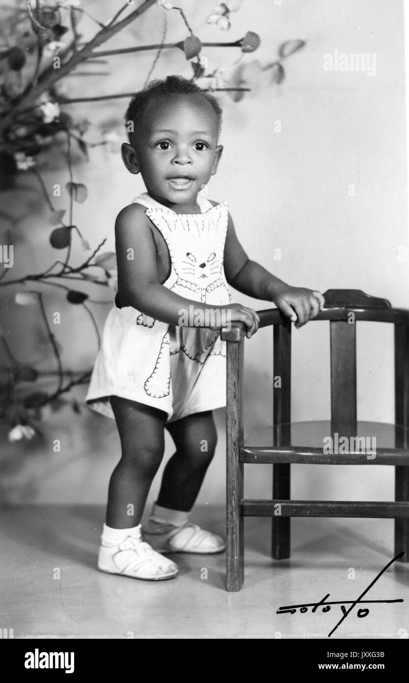 Full length portrait of an African American child, female, wearing white dress, white shoes, holding onto a small chair, smiling facial expression, 1950. Stock Photo
