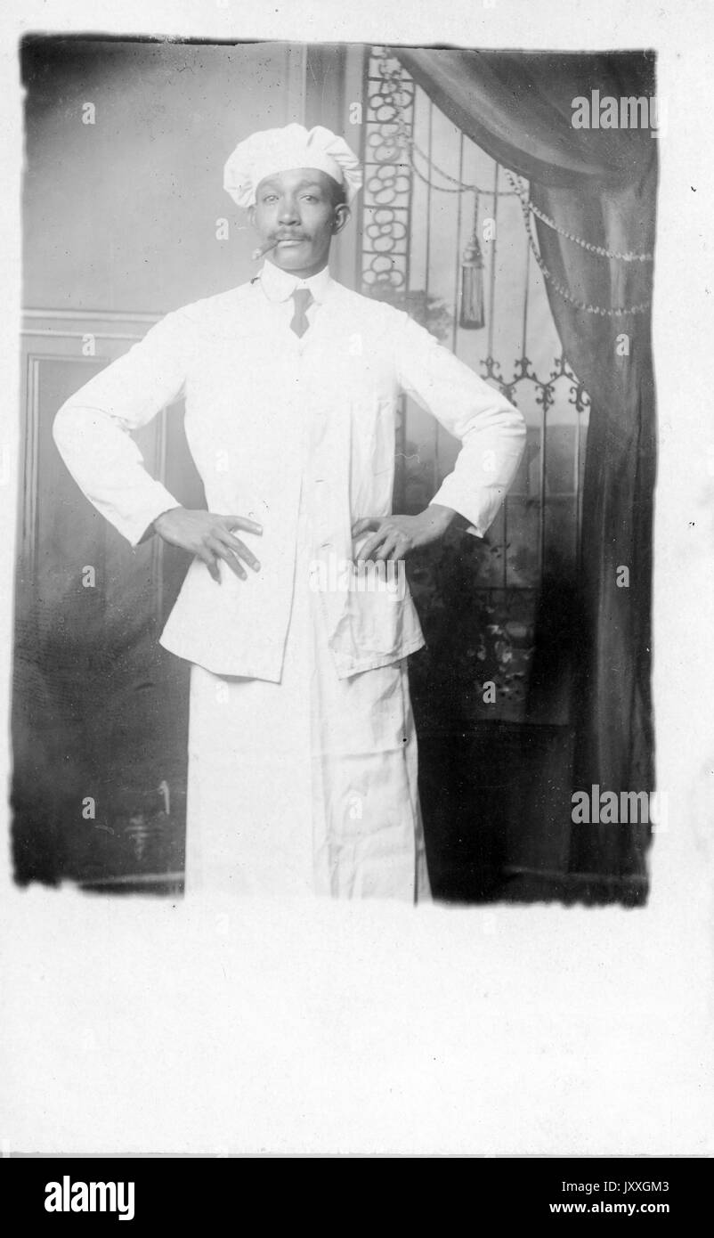 Three quarter length portrait of mature African American man, wearing light outfit, light shirt, dark tie and chef hat, holding cigar in mouth, standing in front of backdrop, neutral expression, 1920. Stock Photo
