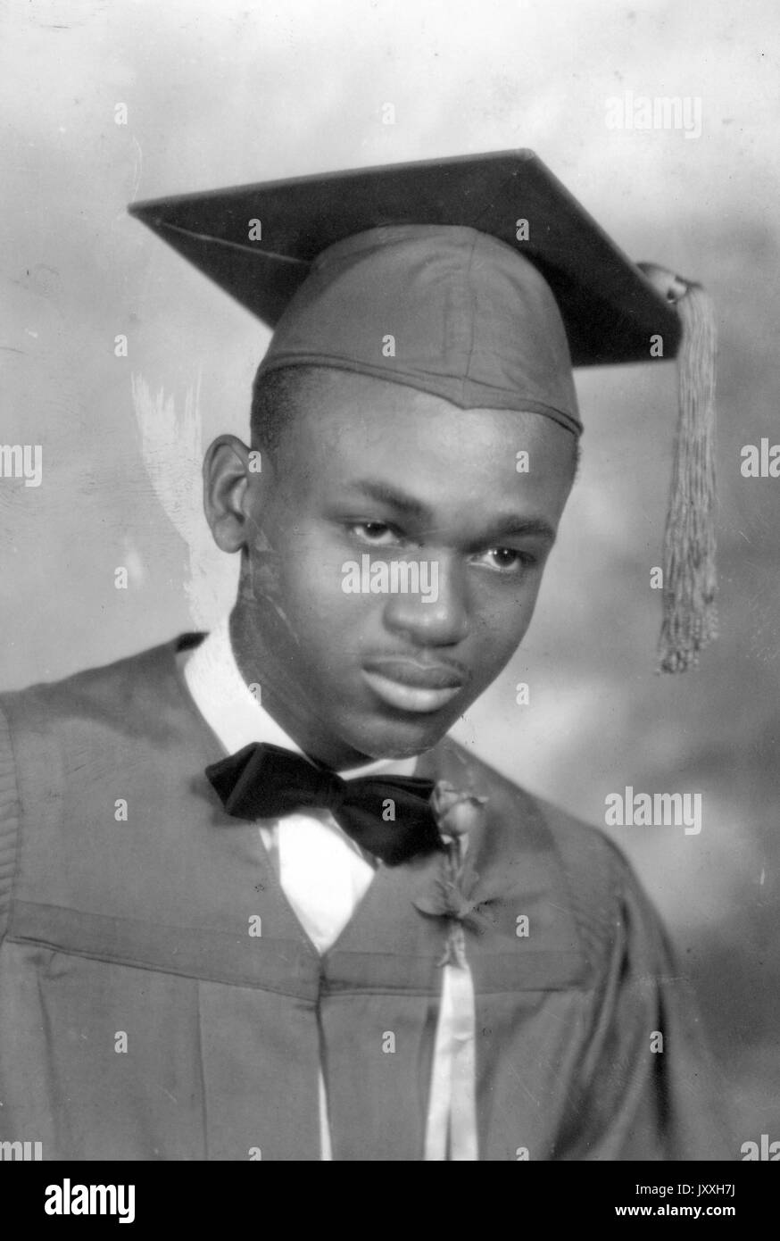 Headshot of young African American man, wearing graduation gown and hat, bow tie, tassel, floral boutonniere and light shirt, neutral expression, 1930. Stock Photo
