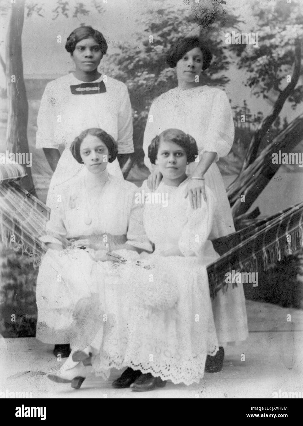 Full length standing and sitting portraits of four young African American women, all wearing light dresses with lace detail, two sitting on hammock, sitting and standing in front of backdrop, neutral expressions, 1920. Stock Photo