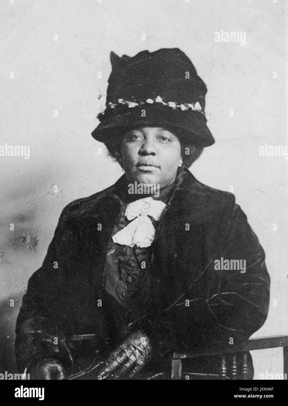 Half length sitting portrait of mature African American woman, wearing dark overcoat, dark blouse with light lace detail, leather gloves and hat, neutral expression, 1920. Stock Photo