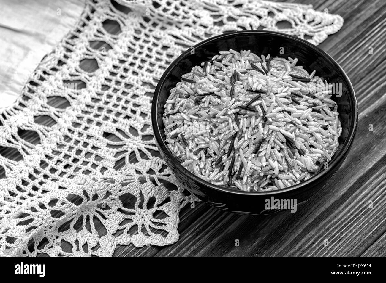 Unpolished brown and black rice in wooden bowl on wooden background with rustic doily crochet. Top view. Black and white Stock Photo