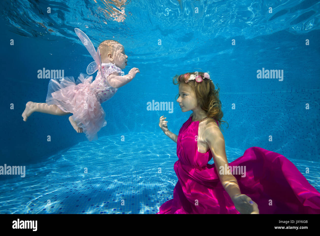 The elder sister in pink dress with little sister in fairy costume posing under water in pool Stock Photo