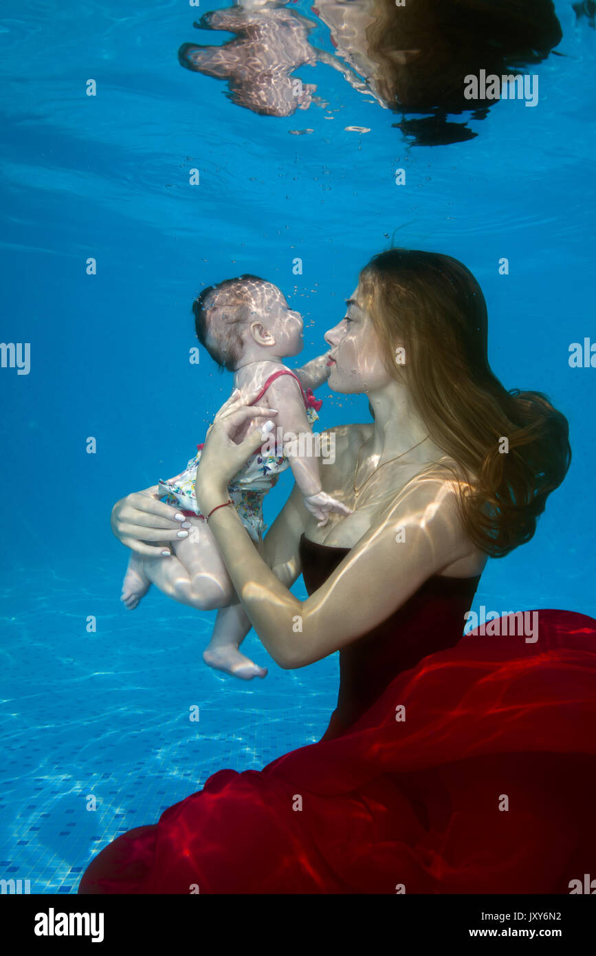 Beautiful young woman in red dress with little girl posing under water in pool Stock Photo