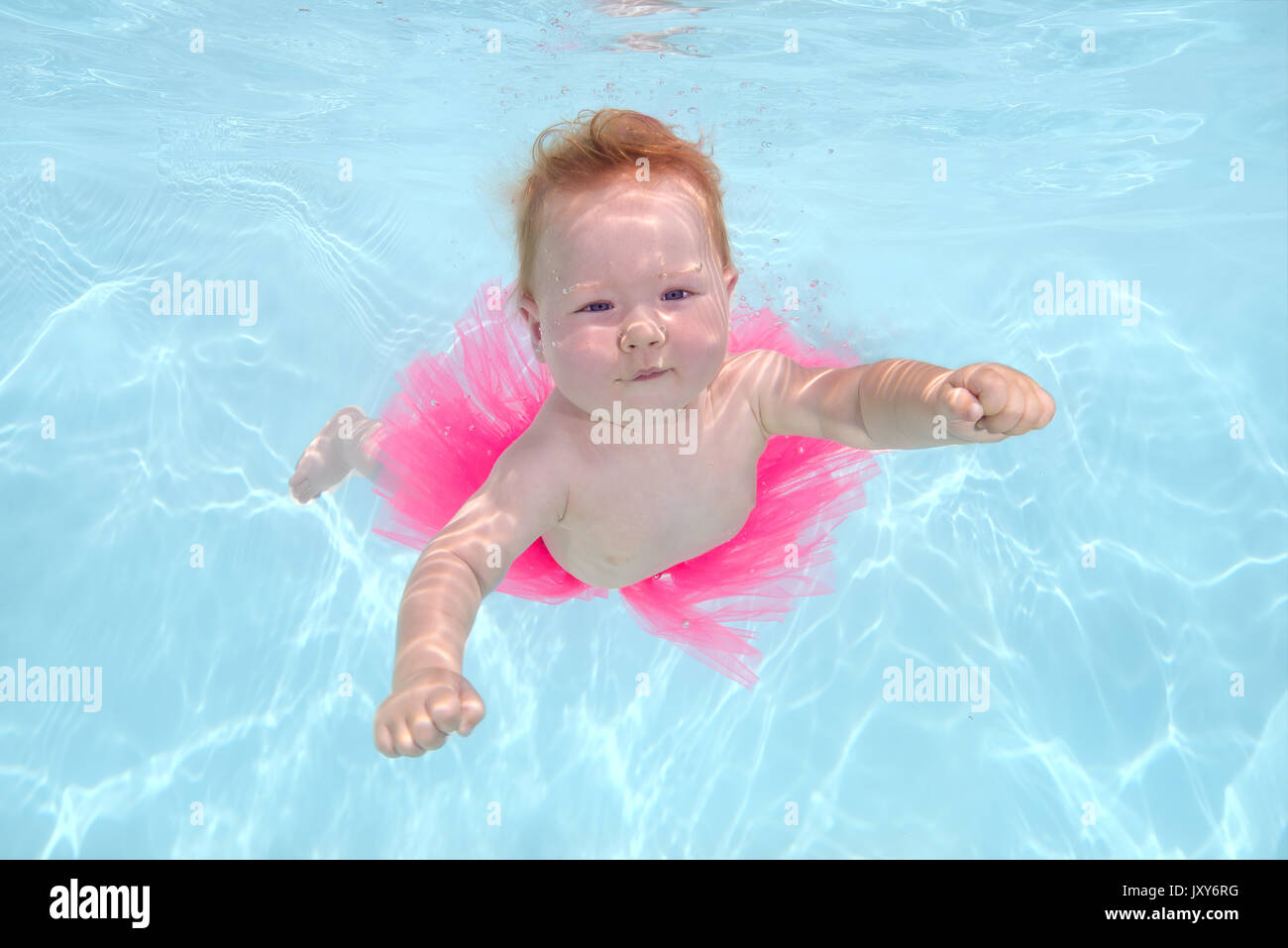 Red-haired little girl in a pink skirt swim under water in the pool Stock Photo