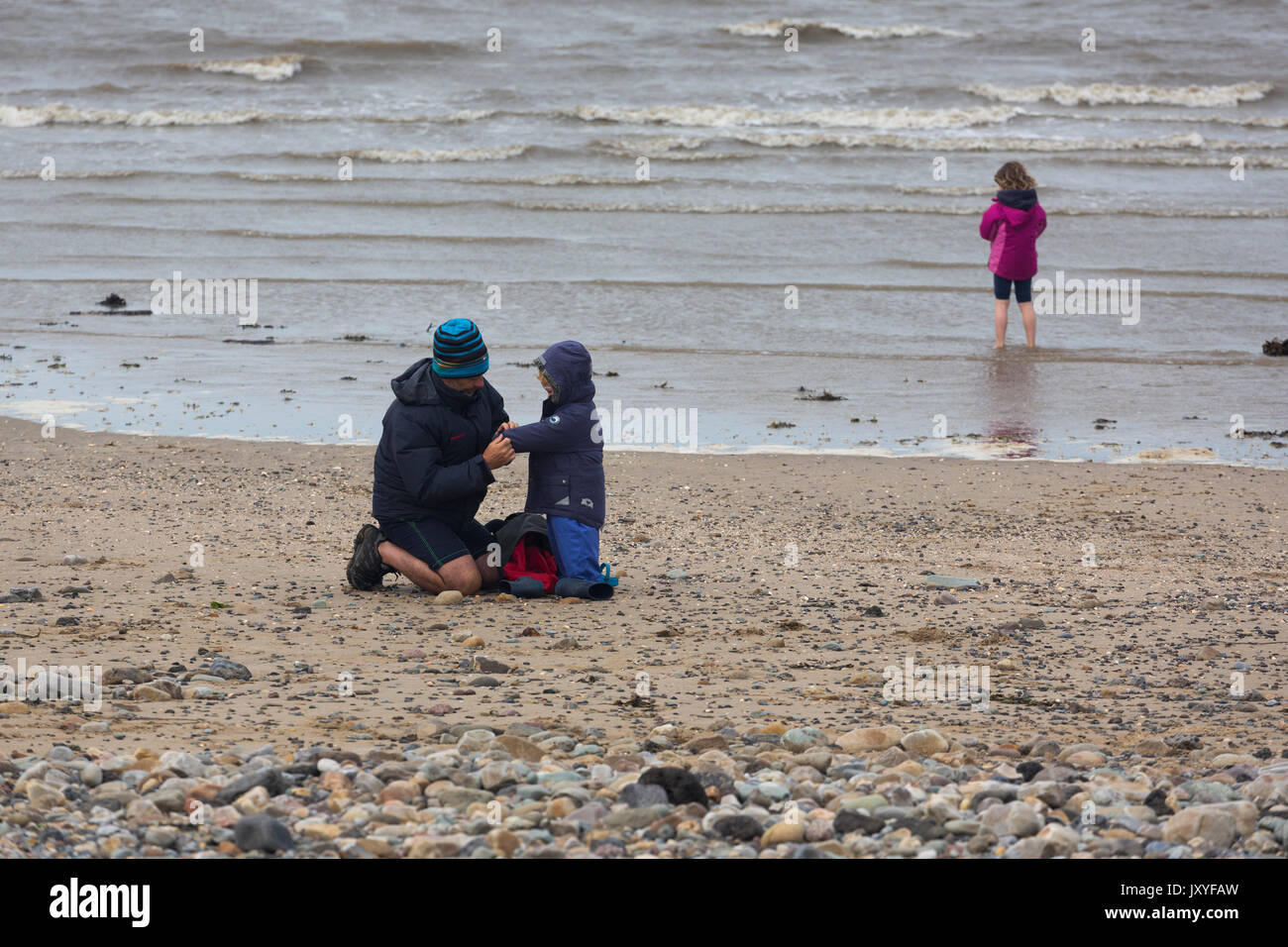 Father and children on beach on cold, wet day with young child paddling in sea Stock Photo