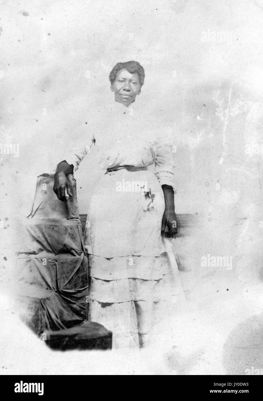 Portrait of an African American woman standing next to a pile of cloth-wrapped items with an upside-down bucket on top, she is wearing a long light colored dress clinched at the waist, her right arm is leaning on the bucket and her left arm is by her side, 1915. Stock Photo
