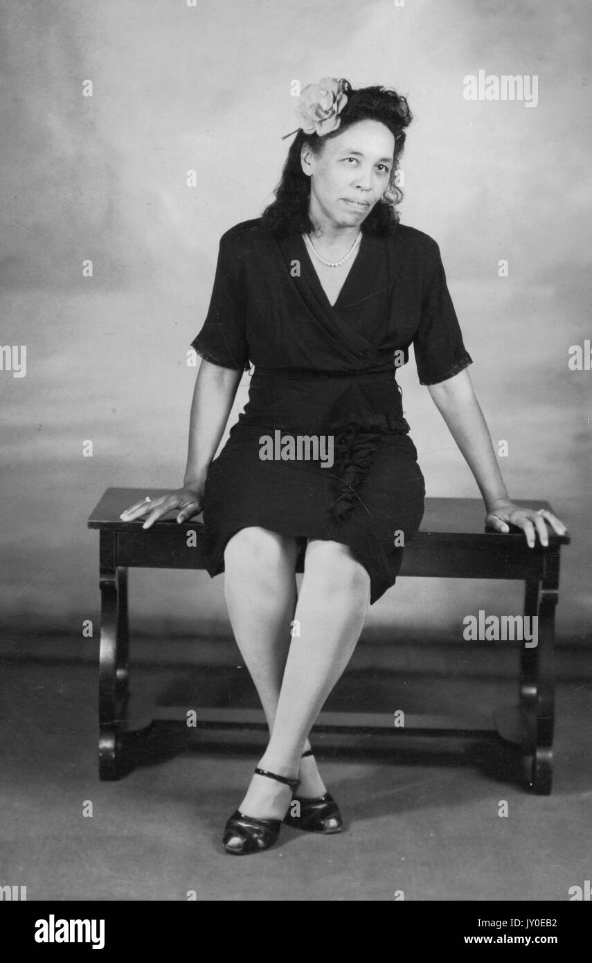 Portrait of an African American woman sitting in front of a mural on a wooden bench, she is wearing a dark colored knee length dress and dark colored heels, she has a flower in her hair and is wearing a pearl necklace, both of her hands are resting on the bench, 1929. Stock Photo