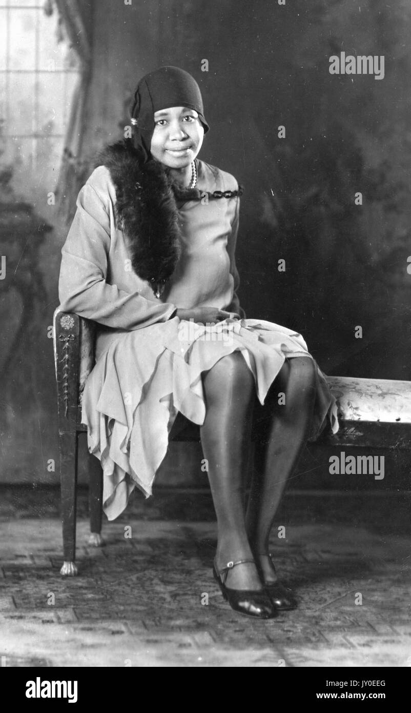 Portrait of an African American woman sitting in front of a mural on an upholstered bench, she is wearing a knee-length skirt and a long-sleeve blouse, she is wearing a stole on one shoulder and a hat, 1929. Stock Photo
