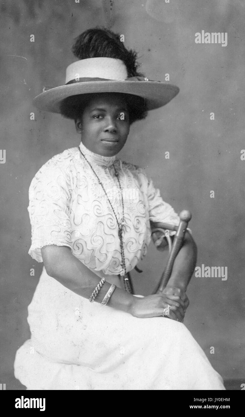 Portrait of an African American woman sitting sideways on a chair, she is wearing a long light colored skirt with a matching blouse, she is wearing a long necklace and a hat, her arms are wrapped around part of the back of the chair and they are resting in her lap, 1929. Stock Photo