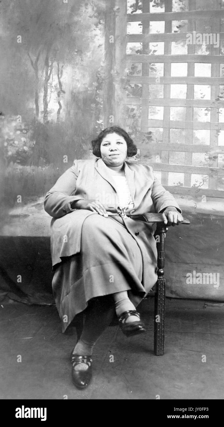 Full length seated portrait of young African American woman with a neutral expression in front of a painted backdrop, holding a book in her left hand, and wearing a lightly colored blouse, skirt, and coat, 1920. Stock Photo