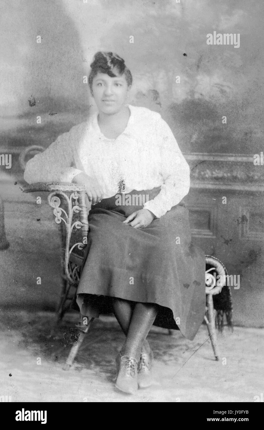 Full length portrait of young African American woman seated in an elaborate chair, wearing a lightly colored blouse, dark skirt, and lace up shoes, 1925. Stock Photo