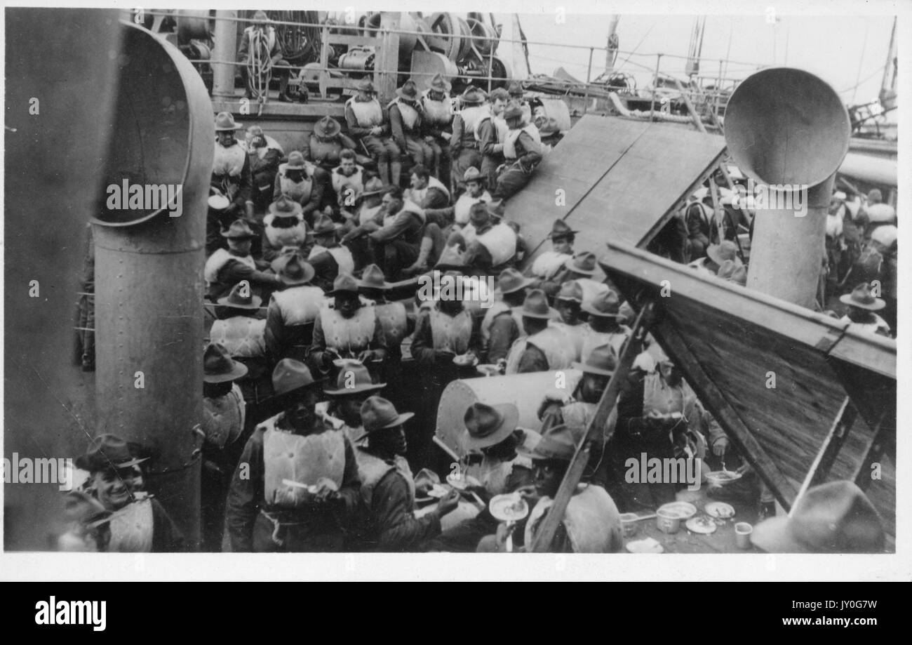 A large group of crew members wearing hats and flotation vests, some of whom are eating, is gathered on the deck of an American vessel, 1915. Stock Photo