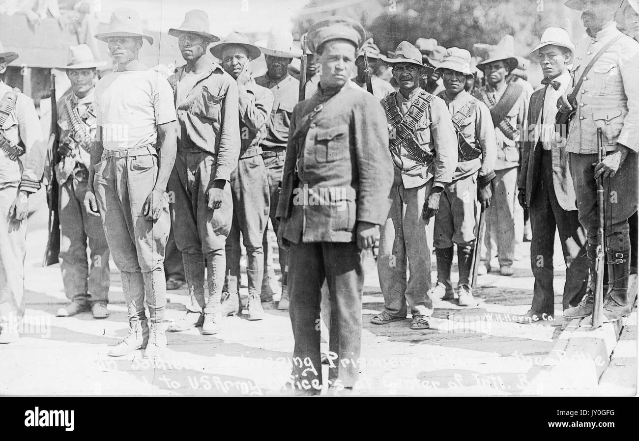 Young and mature African American soldiers with neutral expressions, some of whom carry weaponry, stand in rows outdoors, 1915. Stock Photo