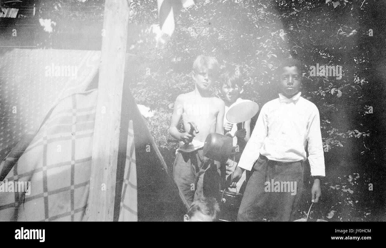 Three quarter length standing portrait of one African American boy with three white boys, African american boy wearing white shirt and dark pants carrying buckets, two white boys wearing pants and holding hammer and tools, standing next to tent, neutral expressions, 1920. Stock Photo