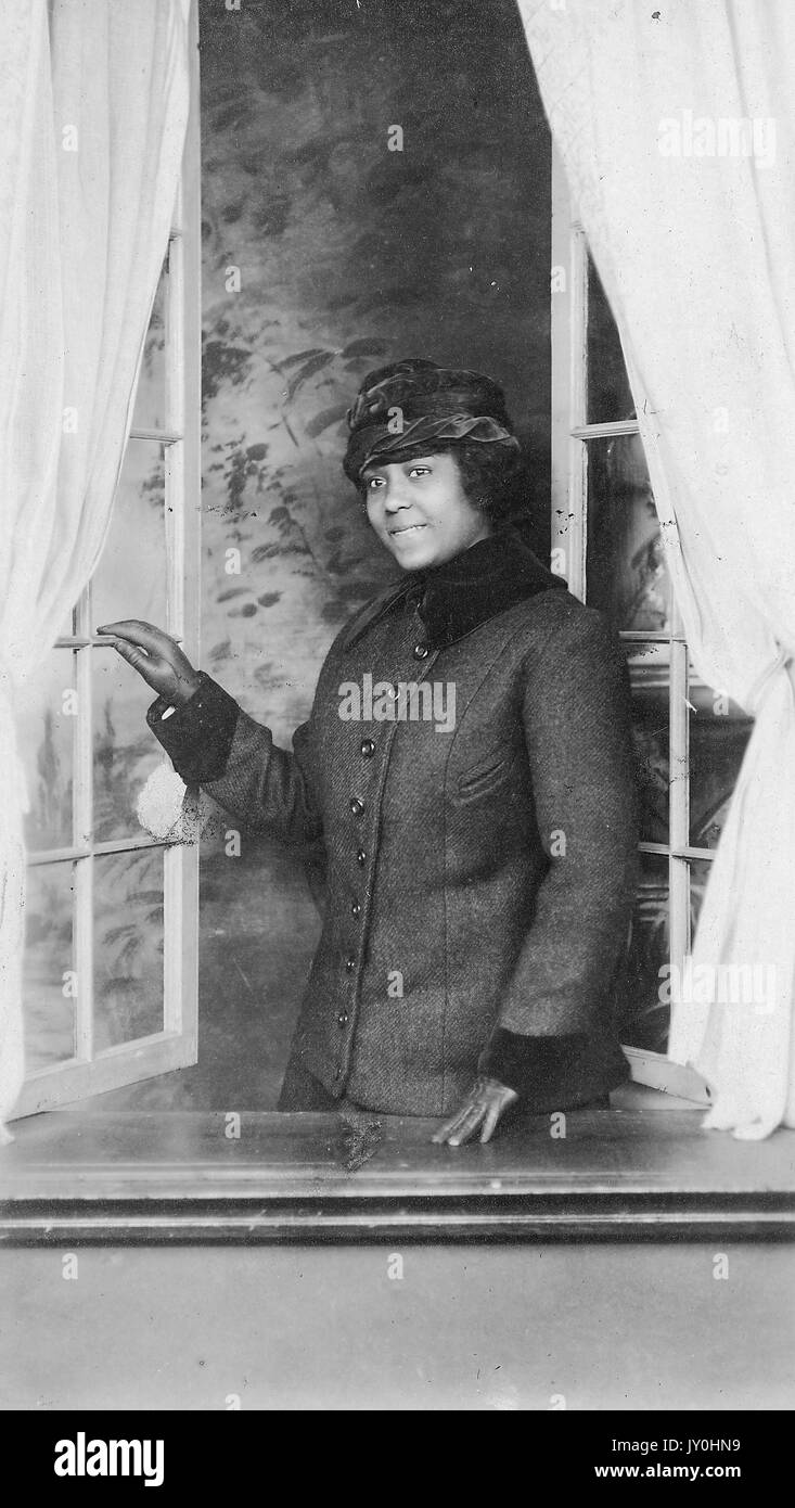 Half length standing portrait of young African American woman, wearing dark overcoat with hat, standing at windowsill while looking outside of open window, smiling expression, 1925. Stock Photo