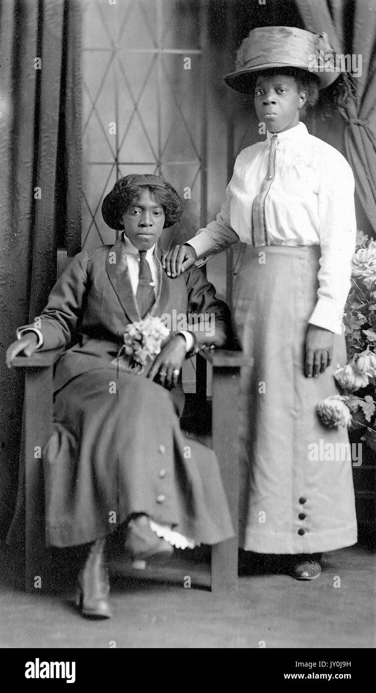 Portrait of two African American woman, one woman is sitting in a large wooden chair on the left, there are some flowers in her lap, she is wearing a long skirt, a suit jacket and a tie, the woman to the right is standing with her right hand on the other woman's shoulder, she is wearing a long skirt, a shirt, a tie, and a large hat, The Davis Studio, 123 W Bay St, Jacksonville, FLA, 1915. Stock Photo