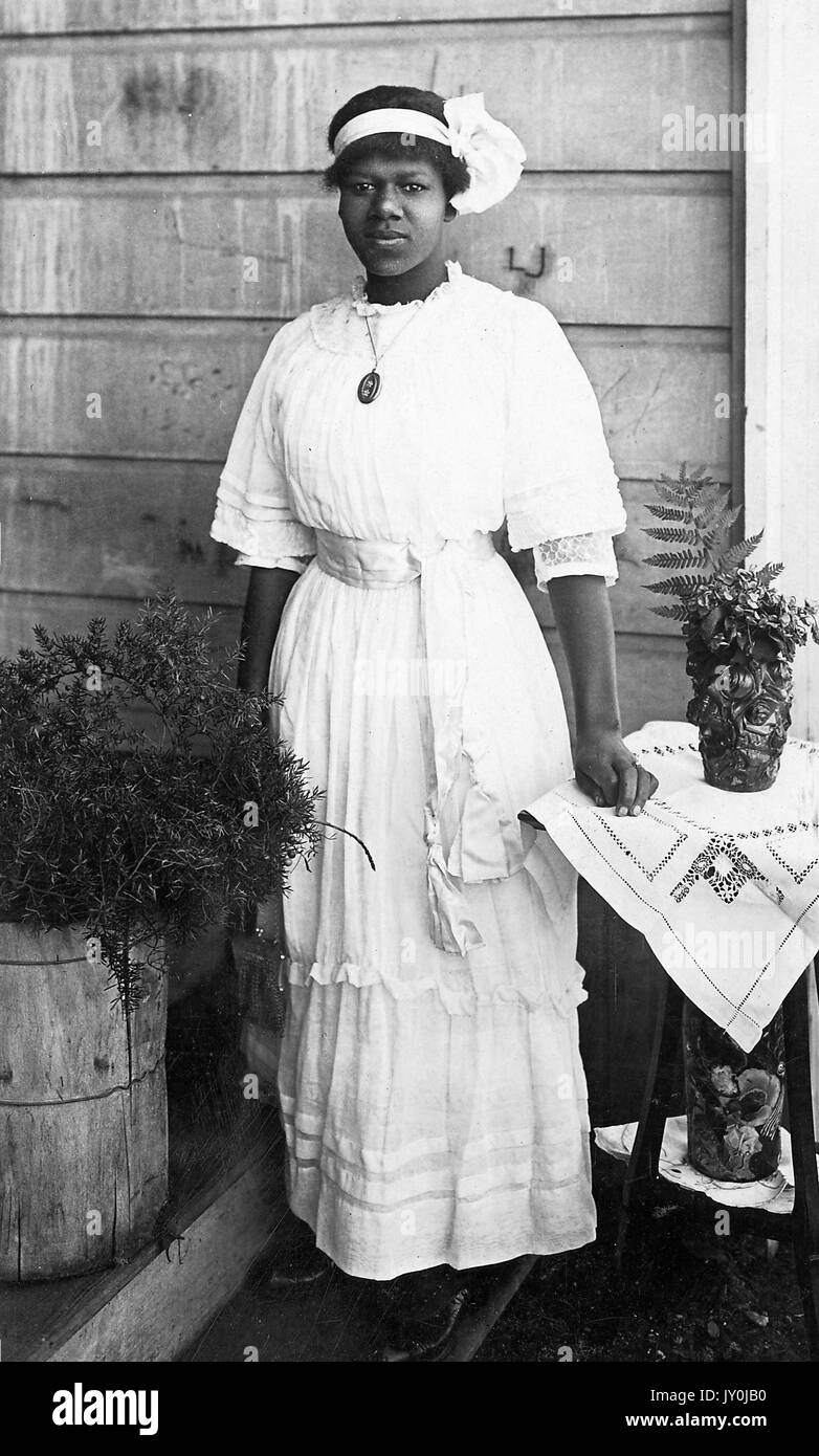 Portrait of an African American woman, she is wearing a long light colored dress and a big pendant necklace, she is also wearing a light colored headband with a large bow or decoration in it, there is a barrel of shrubbery to the left of her and a round wooden table to the left of her, the round wooden table has two levels, there is a flower pot on each level, she is resting her left hand on the round table, 1915. Stock Photo